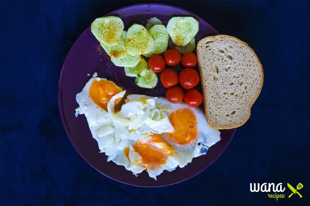 Plate of easy high protein breakfast with fried eggs, sliced cucumbers, cherry tomatoes, and whole-grain bread."