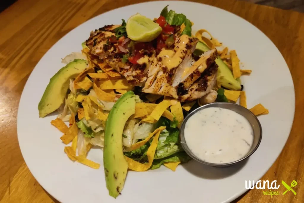 A plated Mexican chicken salad topped with grilled chicken, avocado slices, tortilla strips, and a creamy dressing on the side.